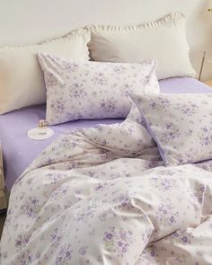 a bed with white and purple sheets and pillows on top of it, next to a night stand