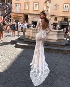 a woman standing in front of a fountain wearing a long sleeved wedding dress with an open back