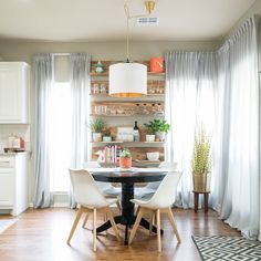 a dining room table surrounded by white chairs and wooden shelves with plants on them in front of the window