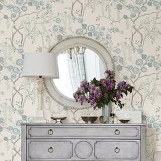 a dresser with flowers on it in front of a wallpapered background and mirror