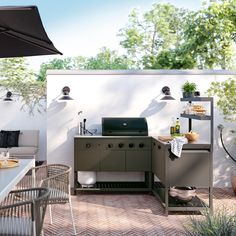 an outdoor kitchen with grill, table and chairs on brick patio next to white wall