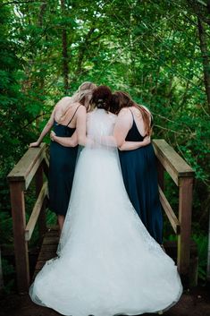 three bridesmaids standing on a wooden bridge in the woods, hugging each other