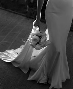 a woman in a long white dress holding a bouquet of flowers and walking down the street