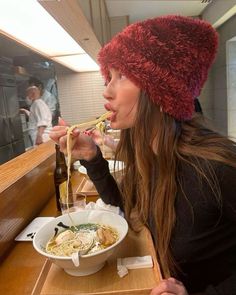 a woman sitting at a table eating noodles with chopsticks in her mouth and wearing a red hat