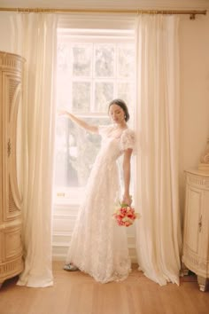 a woman standing in front of a window with her arms stretched out and wearing a white dress