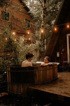 two people sitting in a hot tub with lights strung from the ceiling and trees behind them