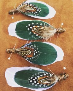 three green and white feathers are sitting on a brown table top with pins in them