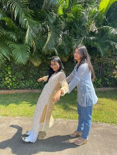 two women standing next to each other in front of some trees and bushes with one pointing at the camera