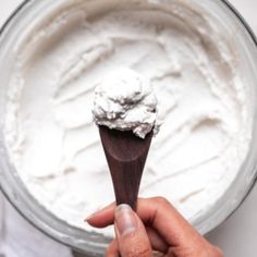 a hand holding a wooden spoon with whipped cream in the bowl next to it on a white surface