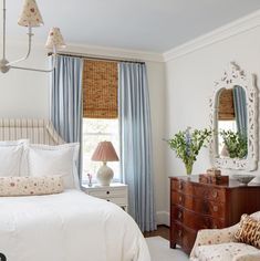 a bedroom with white bedding and blue drapes on the window sill, two nightstands in front of a large mirror