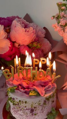 a birthday cake with lit candles on it and flowers in the backgroung