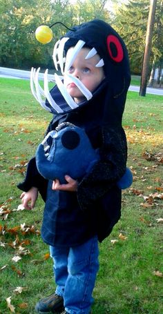 a little boy in a costume standing on the grass with a stuffed animal and tennis ball