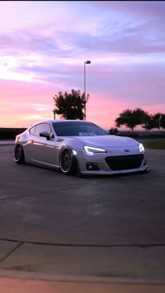 a silver sports car parked in a parking lot at dusk with the sun setting behind it