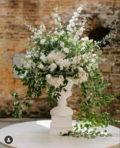 a white vase filled with lots of flowers on top of a table next to a brick wall