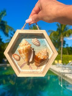 a hand holding a glass ornament with seashells on the beach in front of a swimming pool