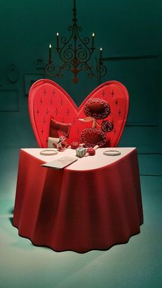 a red heart shaped table with two plates on it and candles in the background, next to a chandelier