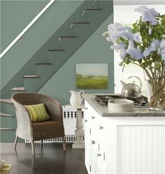 a white kitchen with green walls and brown chairs next to the counter top, has flowers in a vase on it