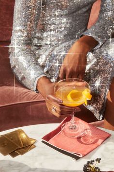 a woman holding a cocktail glass with an orange garnish in it on top of a table