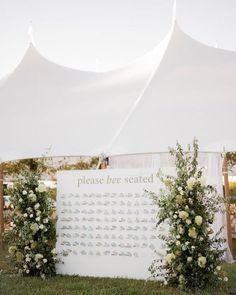 a white tent with flowers and greenery on the grass next to a sign that says please be seated