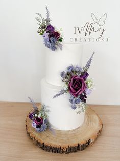 three tiered white cake with purple flowers and greenery on the top, sitting on a tree stump