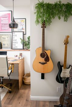 an acoustic guitar is hanging on the wall next to a desk with two guitars and a potted plant