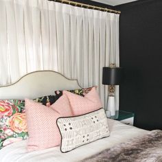 a white bed topped with lots of pillows next to a window covered in sheer curtains