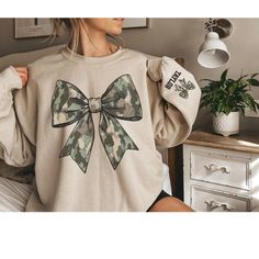 Stay cozy and stylish with our Coquette Camo Sweatshirt, featuring a chic camouflage bow design and personalized sleeve text! 🎀🪖 This trendy sweatshirt is perfect for those who love a blend of fashion and functionality. Whether you're showing off your unique style or supporting someone special, this camo sweatshirt is a must-have for any wardrobe. 🌟 Personalized Just for You: Customize the sleeve with any text of your choice--whether it's your name, a meaningful word, or a special date, make Coquette Sweater, Duck Hunting Shirts, Camo Sweater, Bow Coquette, Camo Shirt, Camo Sweatshirt, Bow Shirt, Womens Camo, Bow Shirts