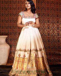 a woman standing in front of a wall wearing a white and yellow dress with gold accents