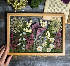 a person holding up a framed photo with flowers and plants in the background on a wooden table