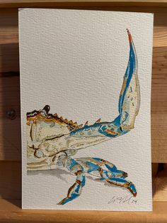 a painting of a blue crab sitting on top of a wooden table next to a piece of paper