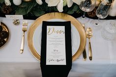 a place setting with white flowers and black napkins, gold rimmed plates and silverware