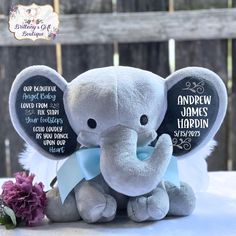 an elephant stuffed animal sitting on top of a table next to a purple flower and plaque