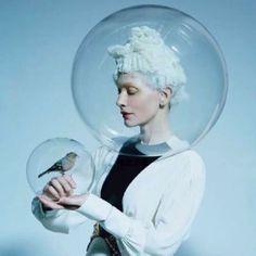 a woman with white hair holding a bird in a glass bowl on her head and wearing a black dress