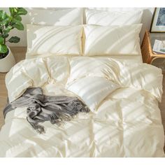 an unmade bed with white sheets and blankets on it, next to a potted plant