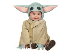 a baby yoda costume sitting on the ground with his hands up and wearing a hat