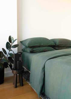 a bed with green sheets and pillows next to a potted plant on the floor