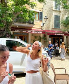 two women eating ice cream in front of a white car and people sitting at tables