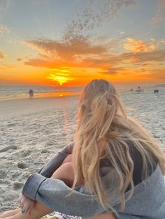 a woman sitting on top of a sandy beach next to the ocean at sun set