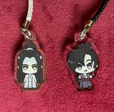 two keychains with anime characters on them sitting on a red cloth covered surface