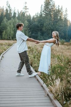 a man and woman holding hands while walking across a wooden walkway in the grass with trees behind them