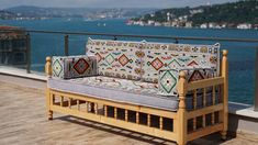 a wooden couch sitting on top of a wooden deck next to the ocean with mountains in the background