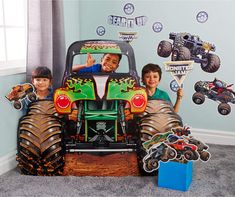 two boys are sitting in the back of a truck with monster trucks on it and another boy is standing behind them