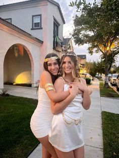 two girls in white dresses hugging each other on the sidewalk near a house and trees