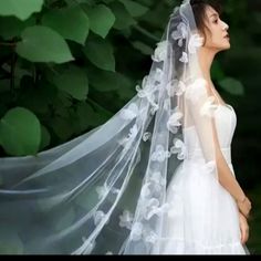 a woman wearing a wedding veil with white flowers on the side is standing in front of green leaves