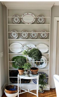 a white shelf filled with plates and potted plants next to a wooden floor in front of a doorway