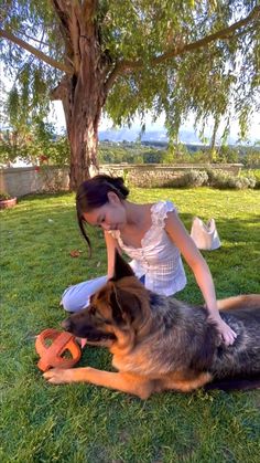 a woman kneeling down next to a dog on top of a lush green field under a tree