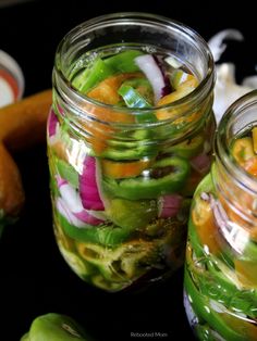 two mason jars filled with pickled vegetables on top of a black table next to carrots and onions