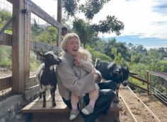 a woman holding a baby and two goats on a bench in front of a fence