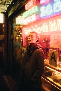 a man standing in front of a store window with neon signs on the building behind him