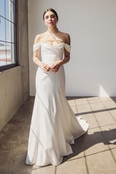 a woman in a white wedding dress standing by a window with her hands on her hips